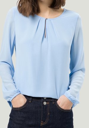 Femme portant un chemisier bleu clair à manches longues avec un petit bouton en forme de trou de serrure, les mains dans les poches d’un jean en denim foncé.