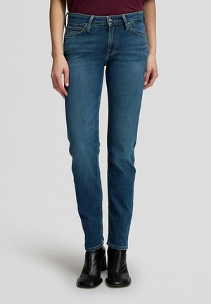 Persona che indossa jeans slim fit blu scuro con tasche frontali e stivaletti neri, in piedi davanti a uno sfondo grigio chiaro uniforme.