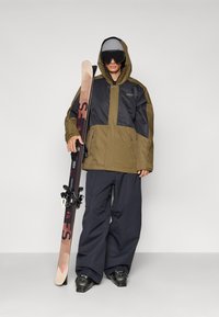 Skioutfit med en hættejakke i olivengrøn og sort med store lommer, parret med løstsiddende marineblå bukser og sorte skistøvler.