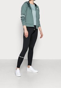 Osoba stojí oblečená v šedé zipové bundě, světle modrém topu, černých legínách a bílých teniskách na jednobarevném pozadí.