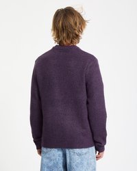 Person med skulderlangt blondt hår iført en mørk lilla striktrøje og lyseblå acid-wash jeans, vender væk mod en hvid baggrund.