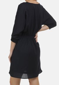 Schwarzes Kleid mit Dreiviertelärmeln, elastischem Bund und einer glatten, leichten Textur. Endet knapp über dem Knie, ohne sichtbare Muster.