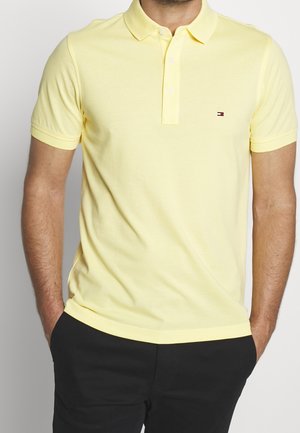 Homme portant un polo jaune clair à manches courtes avec un petit logo sur la poitrine et un pantalon noir, les mains dans les poches.