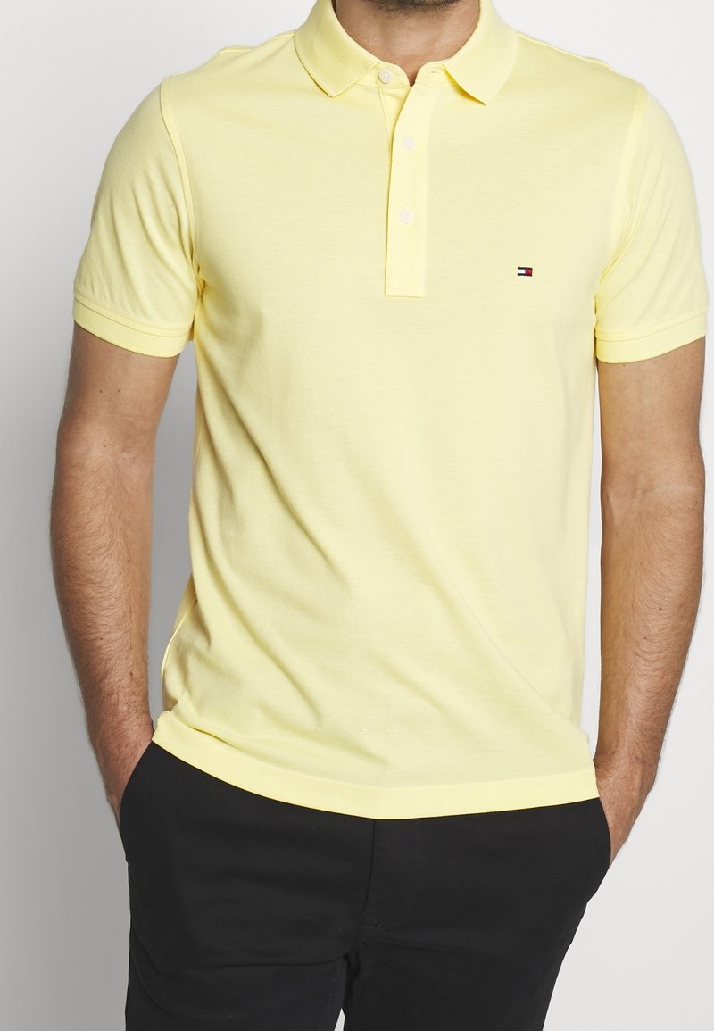 Homme portant un polo jaune clair à manches courtes avec un petit logo sur la poitrine et un pantalon noir, les mains dans les poches.