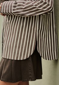 Maglietta a righe marroni e bianche con maniche lunghe, orlo curvo e spacchi laterali, abbinata a una gonna plissettata marrone.