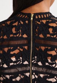 Lähenemine musta pitskleidi seljale, kus on lilleline tikand ja keskel kuldne tõmblukk pruunil nahal.