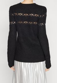 Maglione nero a maglia con maniche lunghe, caratterizzato da un motivo in pizzo sulla schiena. Tessuto testurizzato e polsini a coste. Gonna plissettata argentata visibile sotto.