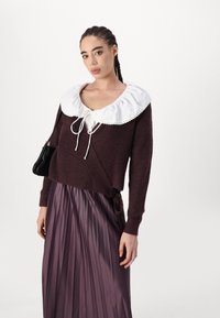 Maglione borgogna con un ampio colletto bianco, legato sul davanti. Abbinato a una gonna plissettata e lucida di colore viola. Una borsa nera completa il look.