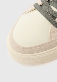 Zapatilla beige con parte superior de cuero liso color crema, cordones texturizados grises y una superposición de ante con textura suave. Suela de goma con estrías.