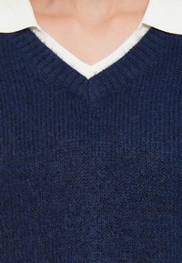 Pulover de culoare albastru marin, cu decolteu în formă de V și textură ribbată, având o cămașă albă cu guler dedesubt. Material tricotat ușor pufoșel.