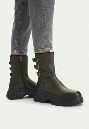 Ciemnozielone skórzane buty do połowy łydki z masywnymi czarnymi podeszwami i trzema bocznymi klamrami, noszone z szarymi dżinsami.
