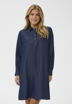 Lächelnde Frau mit langen blonden Haaren, die ein dunkelblaues Jeanshemdkleid mit langen Ärmeln, einem Kragen und Knöpfen auf der Vorderseite trägt.