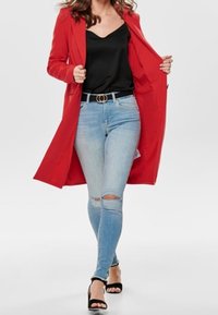 Abrigo largo rojo sobre un top negro, combinado con jeans ajustados de mezclilla clara y desgastados y tacones negros con tiras. Cinturón simple como acento.
