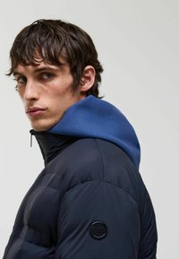 Jachetă puffer de culoare bleumarin, cu un glugă albastră în contrast. Caracteristici o textură matlasată și un patch rotund de logo negru pe mânecă. Închidere cu fermoar.