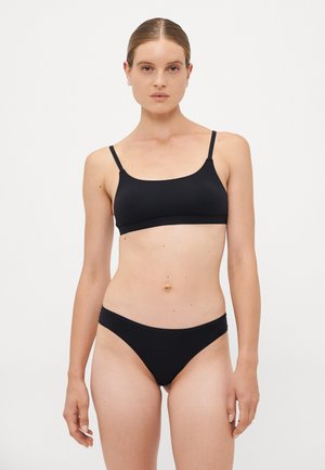 Woman with slicked-back hair wearing a black minimalist bra and matching black underwear against a plain white background.