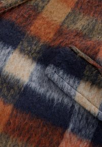 Tissu en laine mélangée à un motif tartan comprenant des teintes orange, marine, crème et marron. Texture douce avec des fibres visibles et un détail de poche.