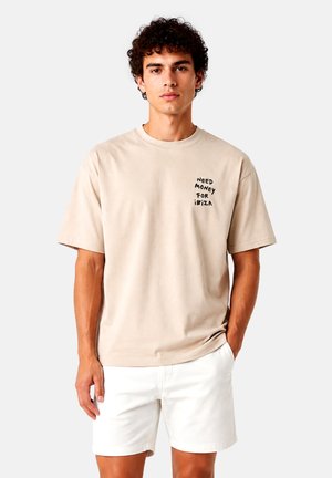 Giovane uomo con capelli ricci che indossa una t-shirt beige con il testo "Need Money for Ibiza" e pantaloncini bianchi, in piedi davanti a uno sfondo semplice.