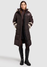 Cappotto lungo imbottito di colore marrone scuro con finitura lucida, dotato di cappuccio e polsini foderati in beige, chiusura con zip e tasche laterali.