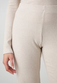 Beige ribstrikkede leggings med høj talje, blød tekstur og tætsiddende design. Nærbillede af stofmønsteret med en hånd hvilende på benet.