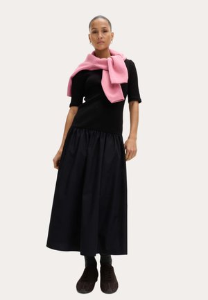 Vrouw met een zwarte jurk met ellebooglange mouwen en een gerimpelde rok, een roze trui over de schouders gedrapeerd, en zwarte schoenen.