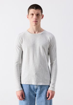 Mladý muž s krátkymi vlasmi, oblečený v jednoduchom svetlosivom tričku s dlhým rukávom a modrých rifliach, stojaci pred bielym pozadím.