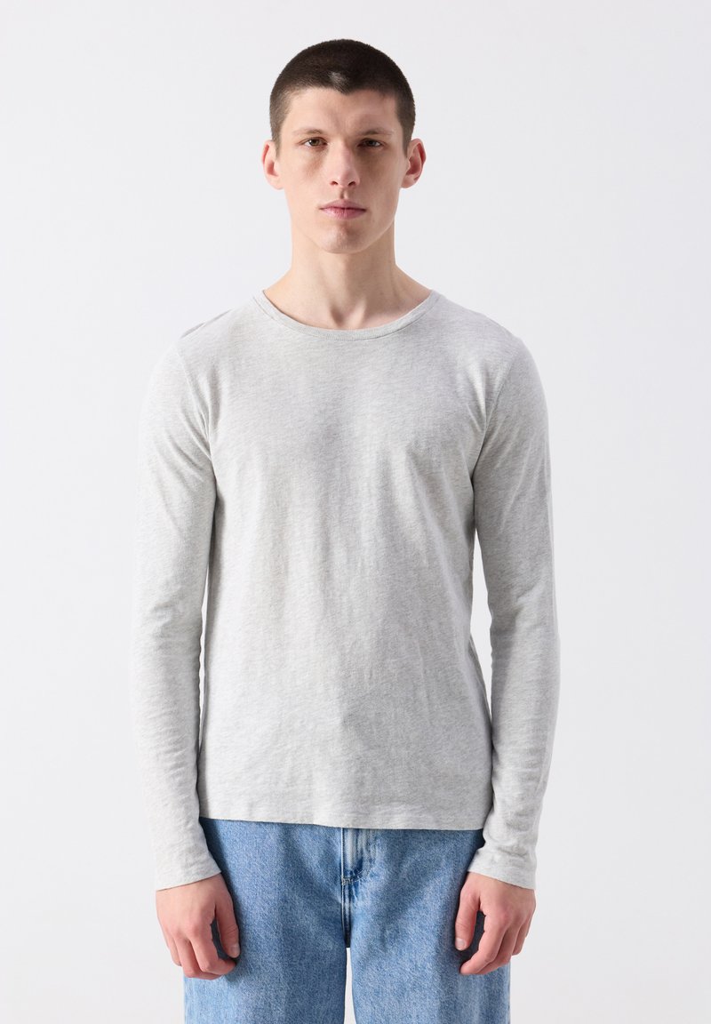 Jonge man met kort haar, gekleed in een effen lichtgrijs shirt met lange mouwen en een blauwe spijkerbroek, staande tegen een witte achtergrond.