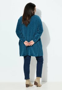 Blå texturerad lång cardigan med en lös passform, i kombination med mörka jeans och beige ankellåga stövlar. Designen har en bred halsringning och långa ärmar.