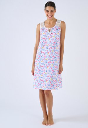 Lächelnde Frau steht barfuß und trägt ein ärmelloses, knielanges weißes Kleid mit rosa, blauem und gelbem Blumenmuster sowie Spitzen-Details an den Schultern.