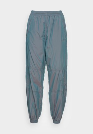 Personne portant un pantalon de survêtement Adidas bleu clair brillant avec des poignets élastiques et des baskets beiges, debout devant un fond blanc uni.