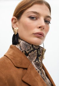 Hnědá semišová bunda s vysokým límcem, se vzorovaným texturovaným zipem. Velké černé a zlaté výrazné náušnice dotvářejí celý vzhled.