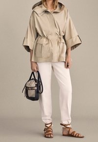 Beżowa kurtka z kapturem z taliowaniem, lekki materiał. Białe spodnie, brązowe sandały na paski oraz czarno-beżowa torebka dopełniają stylizację.