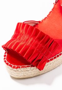 Espadrille roja con una amplia correa de satén con volantes y suela de yute. La parte superior presenta una textura suave, mientras que la suela está trenzada, contrastando con el color vivo.