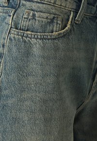 Gai zilu džinsi ar teksturētu virsmu, piecu kabatu dizainu un redzamām šuvēm. Nēsātu izskatu ar nelielu izsistumu malās.