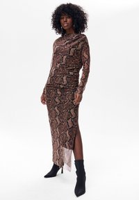 Długa, dopasowana sukienka z materiału w wężowy wzór w odcieniach brązu i czerni, z bocznym rozcięciem i długimi rękawami. Noszona z czarnymi botkami za kostkę.