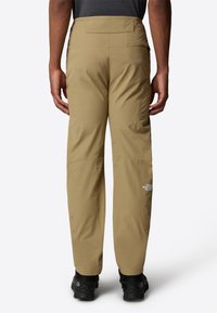 Pantalones de exterior beige con un corte relajado, que cuentan con costuras reforzadas, bolsillos laterales y un detalle de logo en la parte inferior de la pierna. Textura de tela duradera.