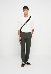 White long-sleeve henley shirt with buttons, olive-green trousers, and black sneakers with brown soles, featuring a black crossbody strap.