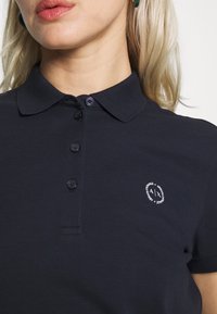 Närbild av en kvinna som bär en marinblå polo med knappkrage och en liten logotyp på bröstet. Ljust hår och diskret smink syns.