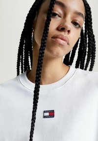 Felpa bianca con scollatura a costine, con un patch del logo "TOMMY JEANS" blu, rosso e bianco sul petto. I capelli intrecciati cadono ai lati.