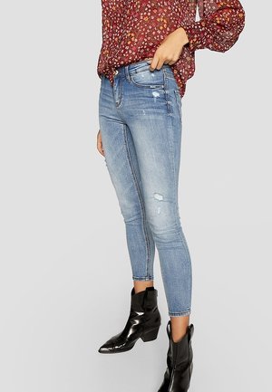 Femme portant un jean skinny bleu clair déchiré, des bottines noires et un chemisier rouge à fleurs à manches longues sur un fond uni.