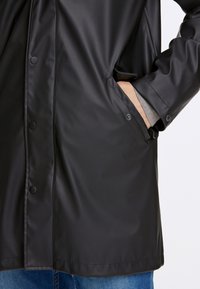 Svart vattentät jacka med tryckknappar, som har fickor på sidan och en slät, glänsande yta. Visas mot blå jeans.