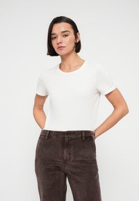 Femme aux cheveux courts et foncés, portant une chemise blanche à manches courtes et un pantalon en velours côtelé marron, debout avec les mains derrière le dos sur un fond uni.
