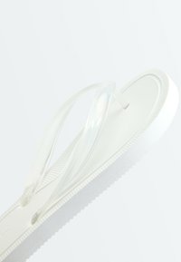 Sandale flip-flop blanche avec semelle texturée et lanières translucides en forme de Y sur un fond clair.