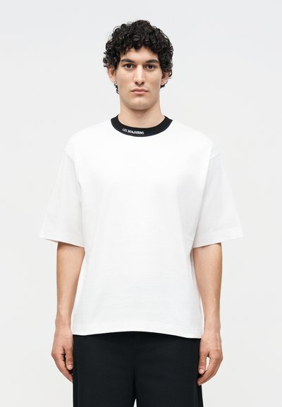 Νέος άνδρας με σγουρά μαλλιά που φοράει μια άσπρη υπερμεγέθη μπλούζα με μαύρο γιακά και το κείμενο "LES BENJAMINS", στέκεται μπροστά σε ένα απλό φόντο.