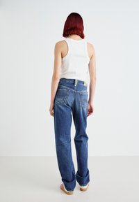 Un paio di jeans in denim a gamba dritta di blu medio, con tasche posteriori con dettagli di cucitura, indossati con una canotta bianca e clogs.
