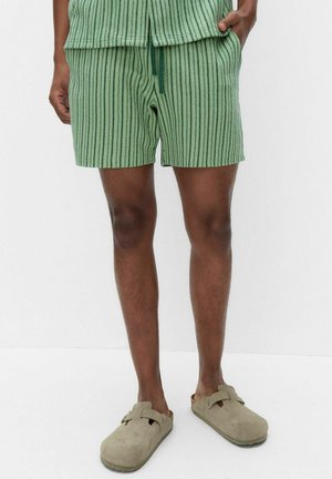 Mann trägt grüne, vertikal gestreifte Shorts und passendes Oberteil, mit beigen Slip-On-Clogs, steht vor einem einfarbigen weißen Hintergrund.