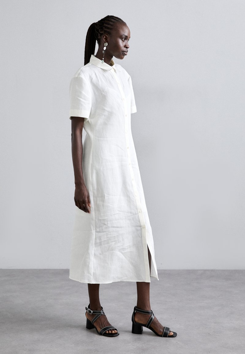 Biała lniana sukienka koszulowa z krótkimi rękawami, z przodu zapinana na guziki i z bocznym rozcięciem. Noszona z czarnymi sandałami na obcasie z cienkimi paskami. Gładka tekstura, minimalistyczny design.