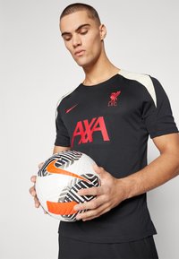 T-shirt de sport noir avec logo rouge L.F.C. et lettering "AXA", présentant des accents beiges sur les épaules. Le modèle tient un ballon de football Nike blanc, noir et orange.