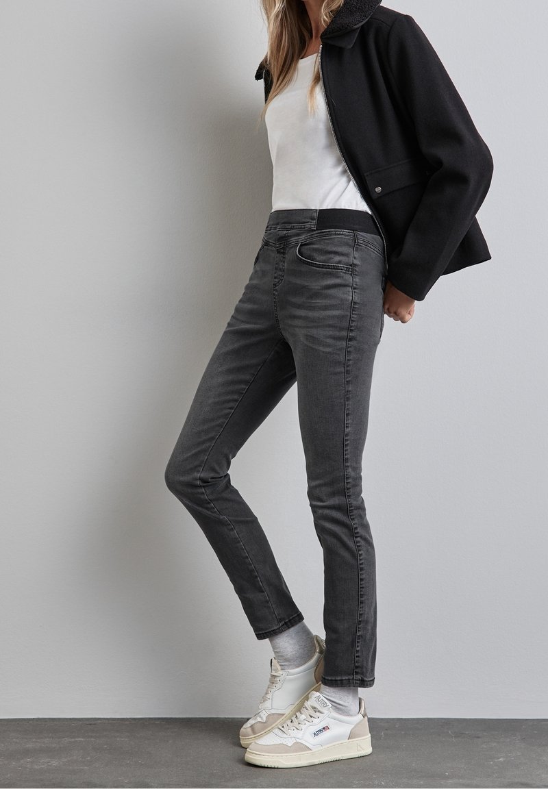 Jean en denim gris coupe slim avec cinq poches, taille mi-haute, texture légèrement décolorée, associés à des baskets blanches et une veste noire.