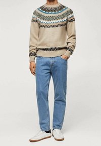 Pull en beige tricoté avec un empiècement à motif en bleu, vert et noir. Un jean bleu clair et des baskets blanches avec des accents marron complètent la tenue.