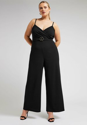 Combinaison noire avec bretelles fines, devant croisé et accent de ceinture circulaire à la taille. Design à jambes larges avec une texture lisse.